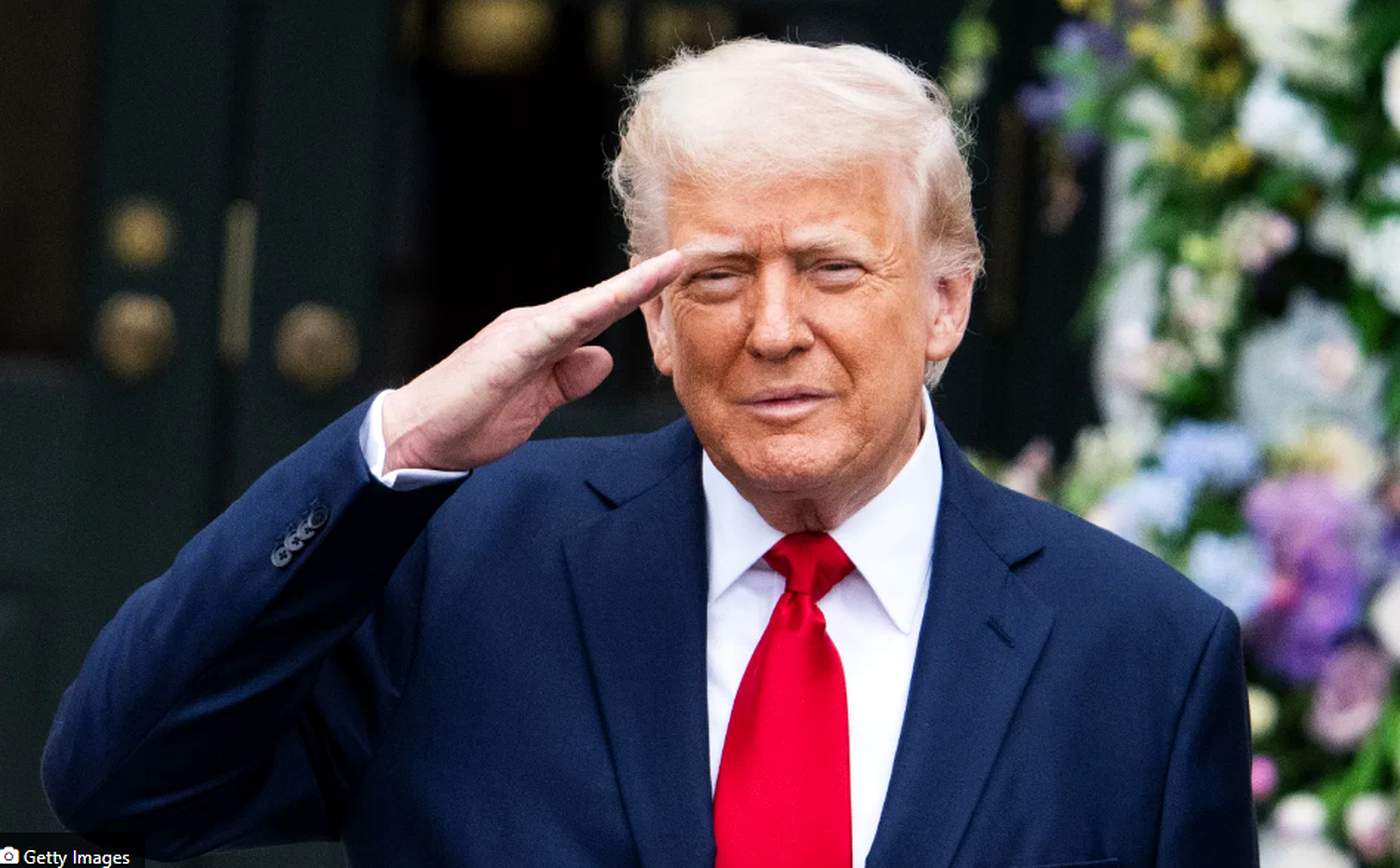 former President Donald Trump salutes outdoors in a dark suit and red tie, with floral